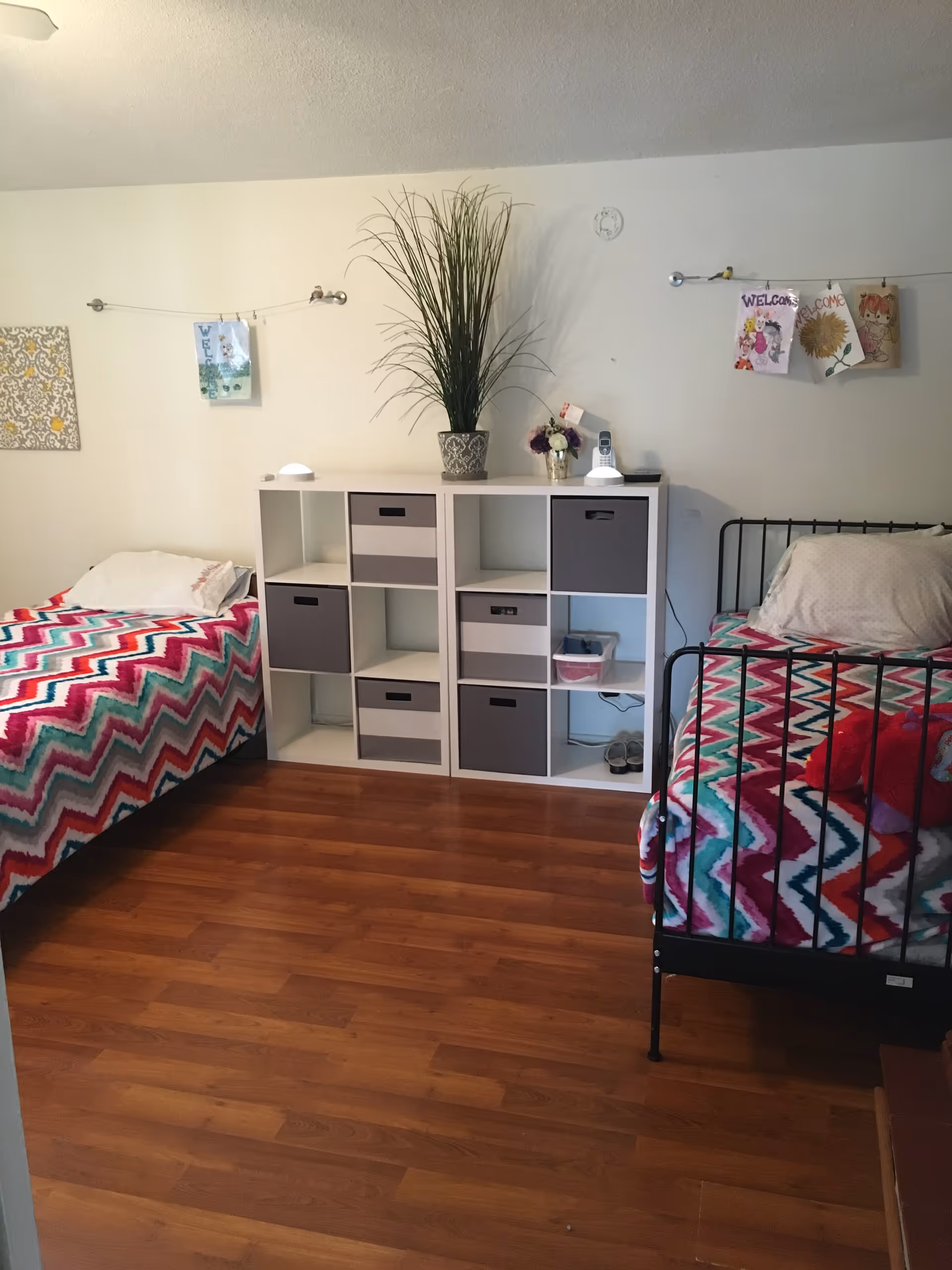 A shared bedroom with two single beds on either side of a white cubical storage unit. Both beds have colorful zigzag patterned blankets in shades of pink, red, teal, and white. The storage unit has several gray and white fabric bins and is topped with a tall potted plant, a small flower arrangement, and a cordless phone. The walls are decorated with children's drawings and welcome signs hung on wires. The floor is wooden.