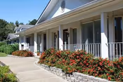 A single story senior living facility exterior with a covered front porch and red flowering shrubs.