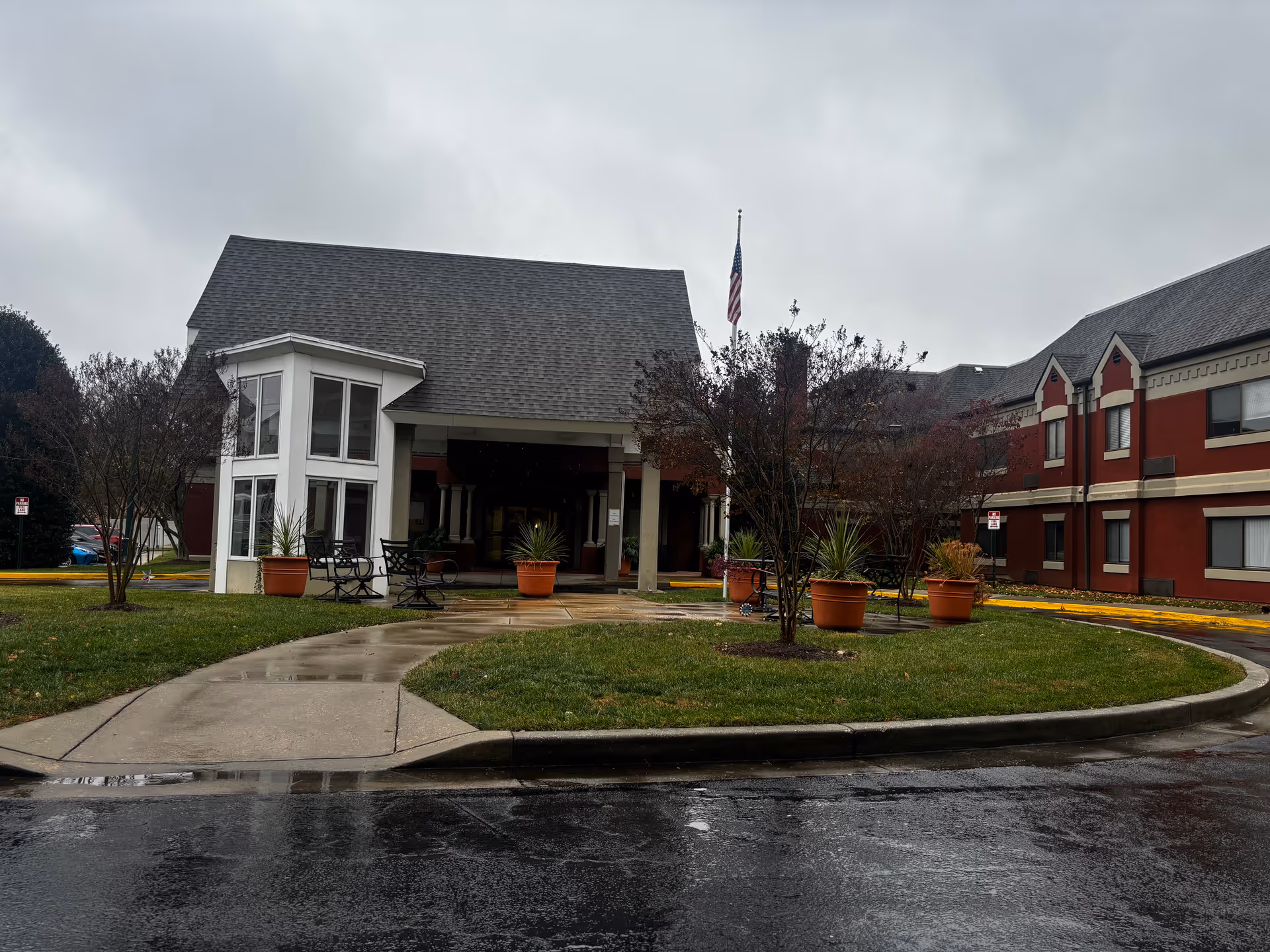 Exterior view of Aspenwood Senior Living Community building on a cloudy day with wet pavement. The entrance features a covered porch with large windows, potted plants, benches, and a flagpole with an American flag. The building is two stories with red and beige walls and a gray shingled roof.