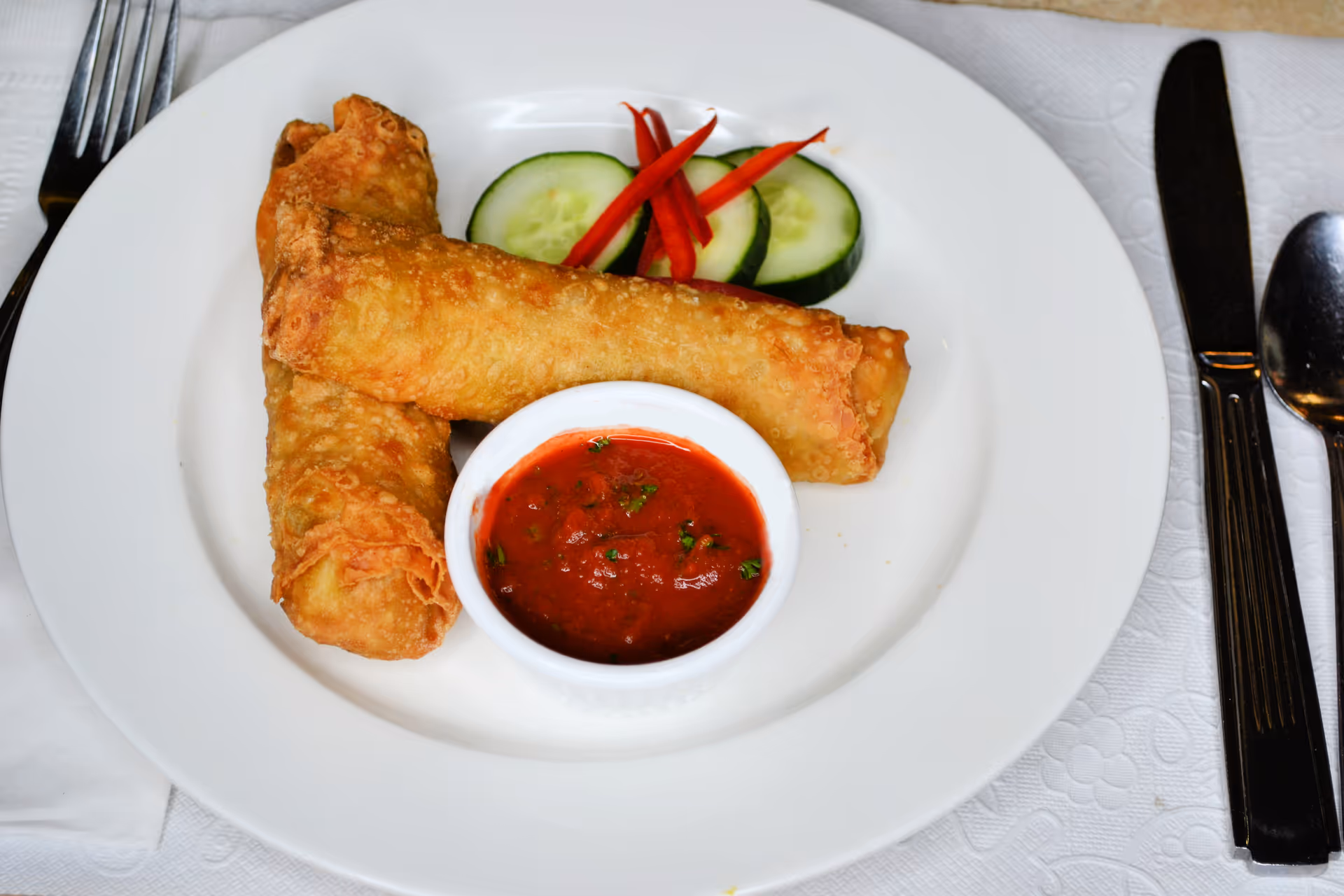 Two fried spring rolls on a white plate with a small bowl of red dipping sauce, cucumber slices and red pepper garnish, with cutlery at the side.