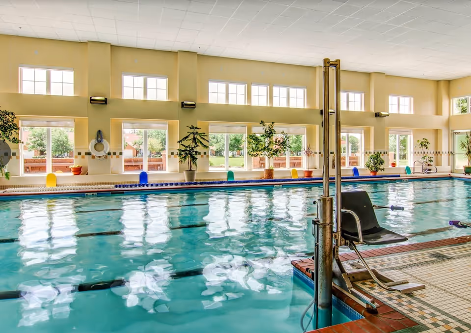 Indoor swimming pool with a mechanical lift, lane markings, large windows and potted plants.