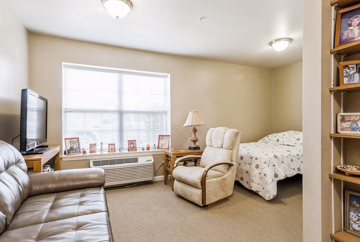 Small studio-style resident room with a bed, recliner, sofa, TV, and a large window with framed photos on the sill.