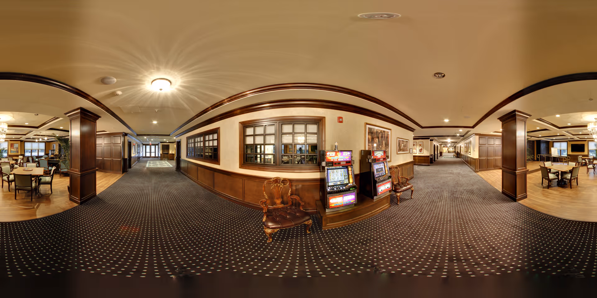 Wide panoramic view of an interior common area in a senior living facility featuring a carpeted hallway with patterned carpet, wooden paneling on the walls, two slot machines, and several seating areas with tables and chairs. The space is warmly lit with ceiling lights and has framed artwork on the walls.