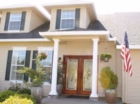 Front entrance of a house with double decorative glass doors, white columns, hanging plants, and an American flag.