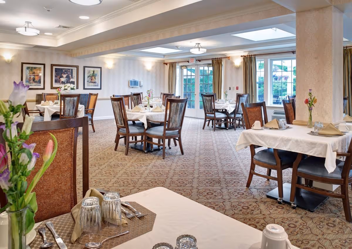 A bright and elegant dining room in a senior living facility with multiple tables set with white tablecloths, napkins, glasses, and silverware. The room features patterned carpet, framed artwork on the walls, large windows with curtains, and ceiling lights. Fresh flowers in vases decorate the tables.