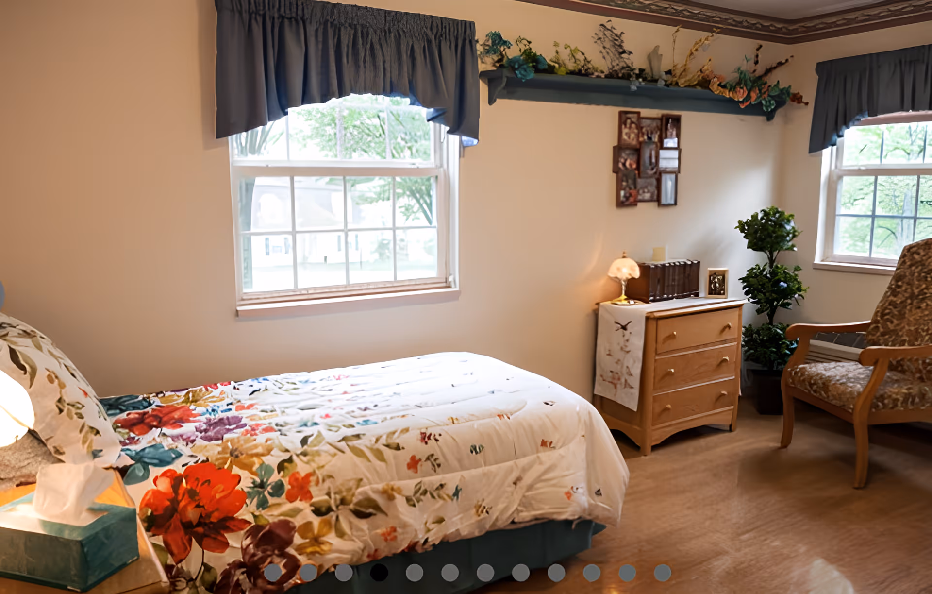 A cozy bedroom with a bed covered in a floral patterned comforter, two windows with blue valance curtains, a wooden dresser with a small lamp and decorative items, a framed photo collage on the wall, a potted plant, and a cushioned armchair.