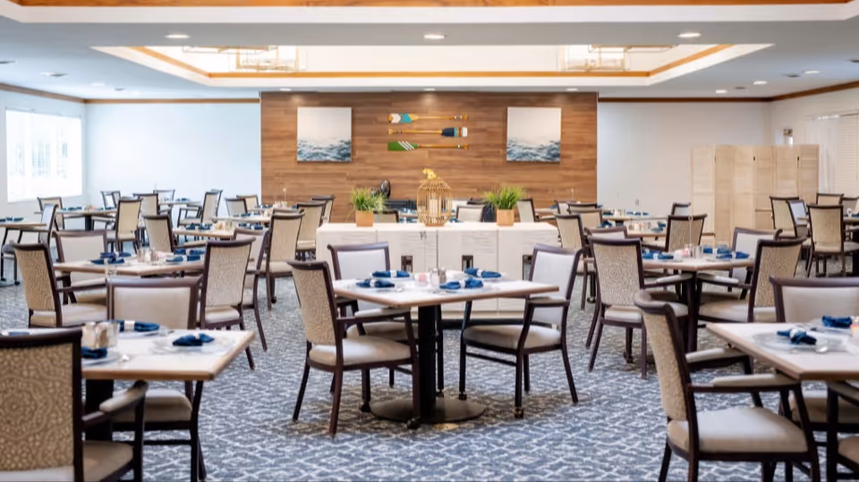 A spacious dining room with multiple tables and chairs arranged neatly. Each table is set with blue napkins, plates, and silverware. The room features a patterned carpet, large windows letting in natural light, and a wooden accent wall decorated with two paintings and three decorative oars. There are also plants and a decorative birdcage on a central table.