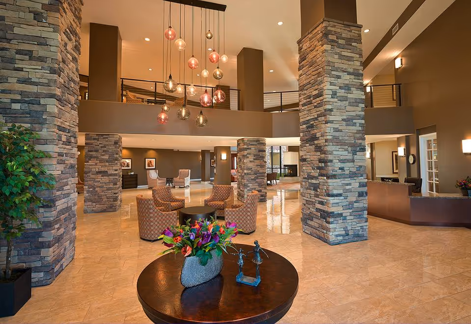 Spacious and well-lit assisted living facility lobby with stone pillars, modern hanging light fixtures, comfortable seating areas with patterned chairs, a round table with a flower arrangement and decorative sculpture, and a reception desk in the background.