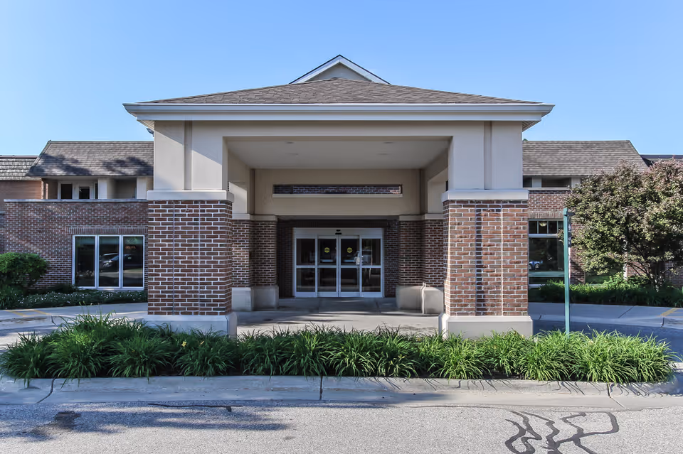 Front entrance of a brick building with a covered driveway and glass double doors, surrounded by greenery and a clear blue sky.