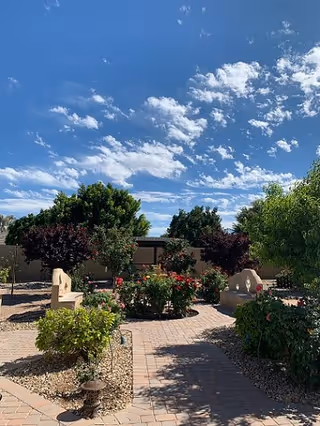 Outdoor garden area with paved walkways, green bushes, flowering plants, and several trees under a blue sky with scattered clouds. Two stone benches are positioned along the path.