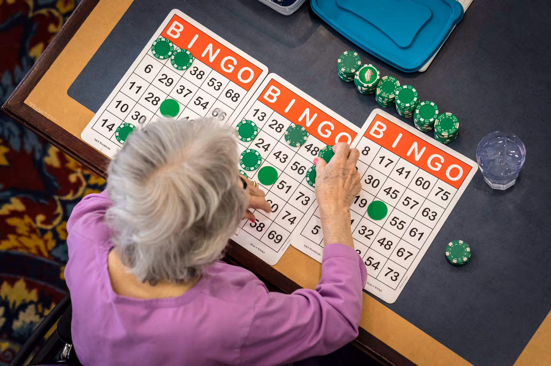 Overhead view of an elderly person with gray hair wearing a purple shirt playing bingo at a table. Three bingo cards with green chips placed on various numbers are visible, along with a stack of green poker chips and a glass of water on the table.