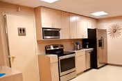 A clean and modern kitchen area with light wood cabinets, a stainless steel microwave, stove, and refrigerator. There is a door with a restroom sign on it to the left, and a decorative sunburst wall clock on the right wall.