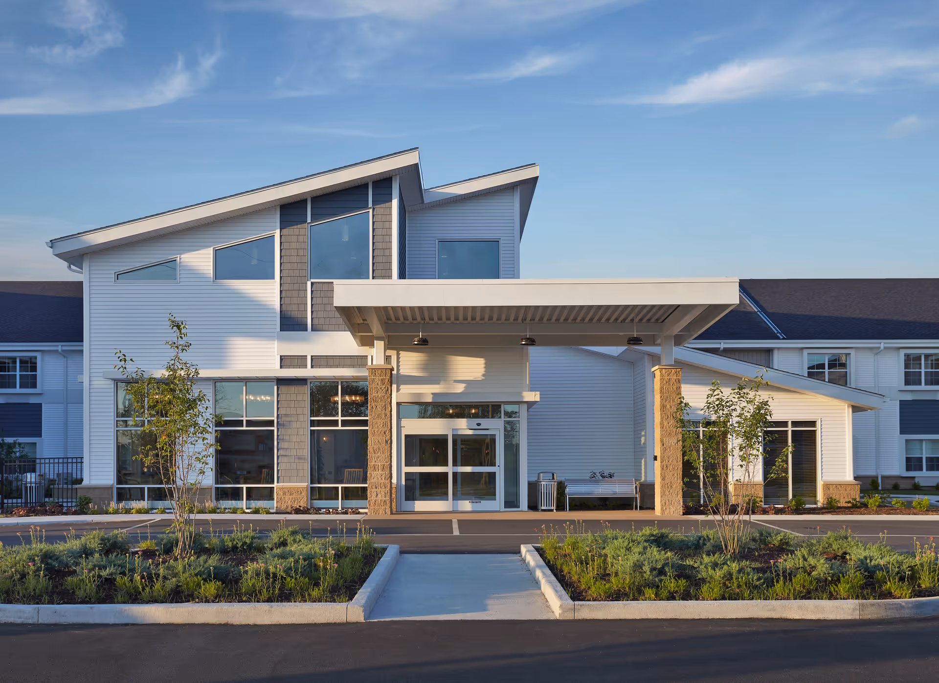 Front exterior view of Harrison's Crossing Health Campus building with modern architecture, large windows, a covered entrance supported by stone pillars, landscaped greenery, and a clear blue sky.