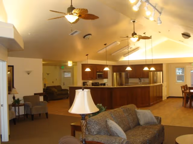 Interior view of a senior living facility showing a spacious common area with a kitchen island, wooden cabinets, stainless steel appliances, ceiling fans with lights, a sofa with cushions, armchairs, and a dining table with chairs in the background.