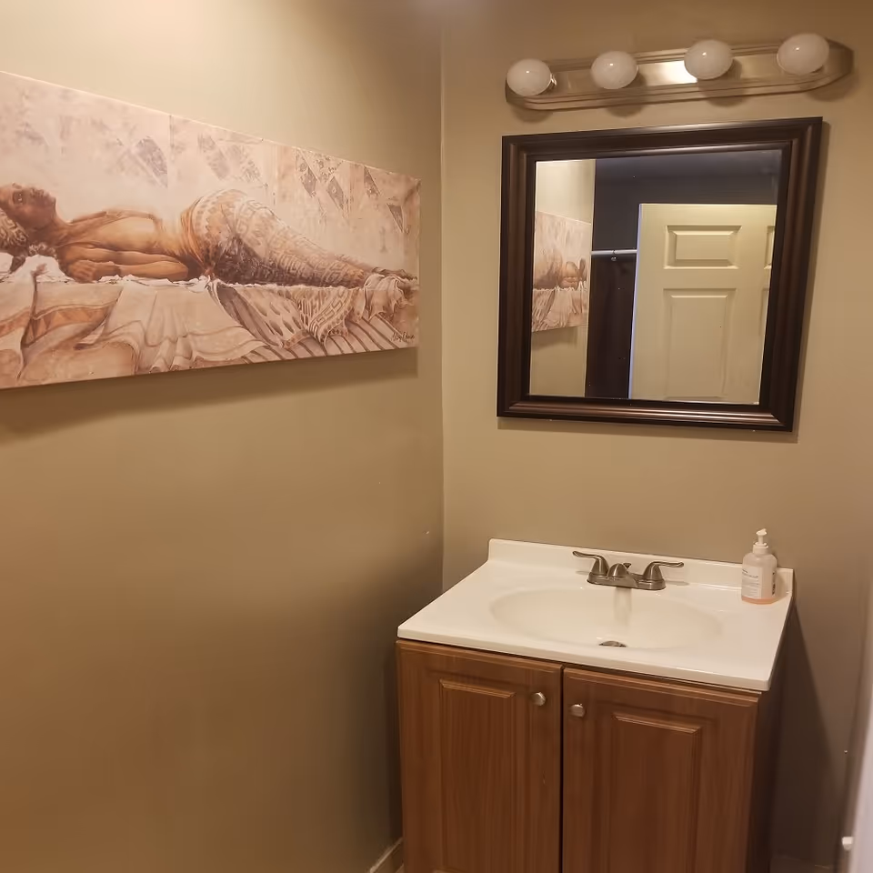 A small bathroom vanity area with a white sink and wooden cabinet below. Above the sink is a rectangular mirror with a dark brown frame and a light fixture with four round bulbs. On the wall to the left of the mirror is a horizontal painting of a reclining woman with tribal tattoos. A door is partially visible in the mirror's reflection.