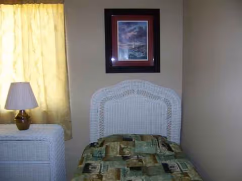 A small bedroom with a white wicker bed, matching dresser, lamp, and yellow-curtained window.