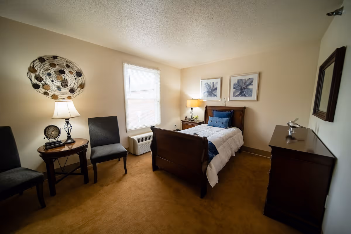 A cozy bedroom with a single wooden bed dressed in white and blue bedding, two framed floral artworks on the wall above the bed, a wooden nightstand with a lamp and small decor, two gray chairs with a round wooden side table holding a lamp and a clock, a window with blinds, and a wooden dresser with a decorative item on top.