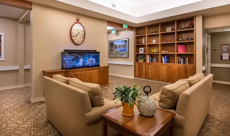A cozy seating area in a senior living facility with two beige sofas facing each other around a wooden coffee table holding a potted plant and decorative item. Behind one sofa is a wooden cabinet with a large aquarium on top and a round wall clock above it. In the background, there is a large wooden bookshelf filled with books and decorative items, and a framed picture on the wall labeled 'THE LAKES'. The space is carpeted with patterned flooring and has soft, warm lighting.