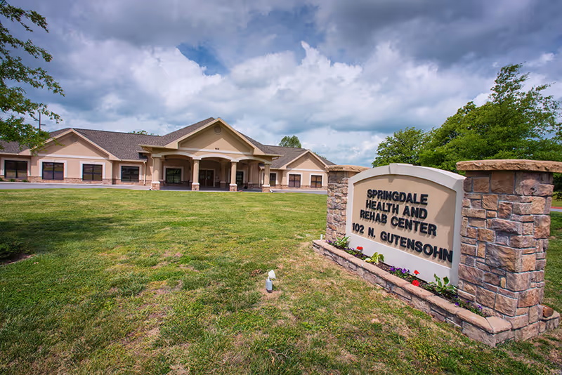 Front exterior of Springdale Health and Rehabilitation Center with a stone entrance sign on a grassy lawn and the single-story building under a cloudy sky.