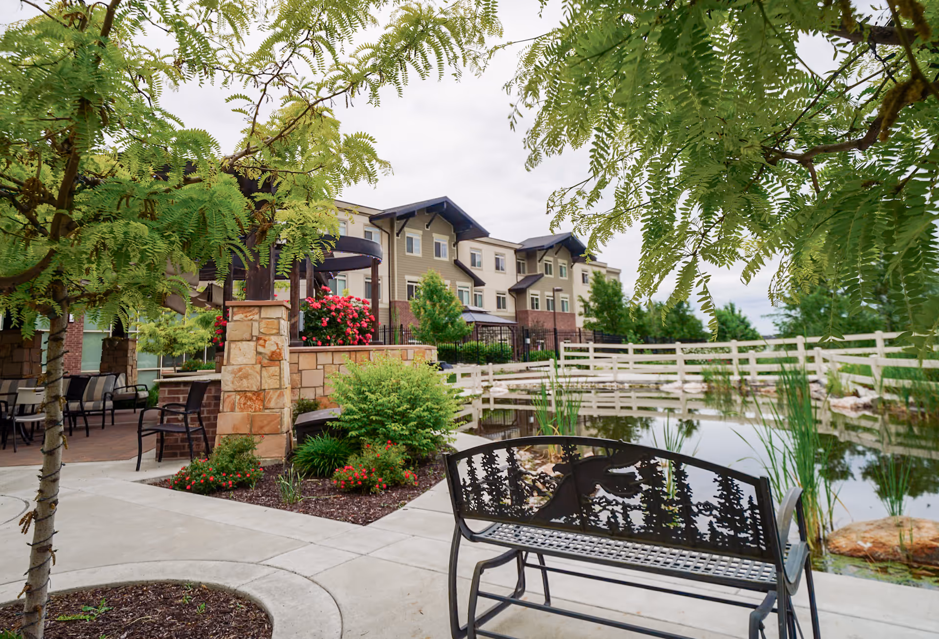 Outdoor courtyard of a senior living facility with a pond, metal bench, landscaped plantings, and the building in the background.