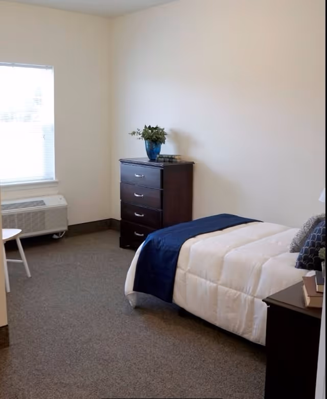 A simple bedroom with a single bed covered in a white comforter and a dark blue throw blanket. There is a dark wooden dresser with a blue vase and some books on top. A window with blinds is on the left side, and a small white chair is partially visible near the window. The walls are plain and light-colored, and the floor is carpeted.