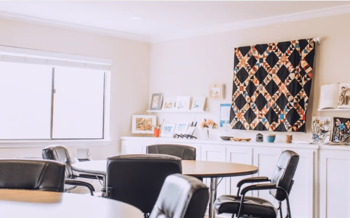 A bright room with round tables and black leather chairs arranged around them. The room has a large window with white blinds, white walls, and a decorative quilt hanging on the wall. Shelves and cabinets along the wall display framed pictures, books, and small decorative items.