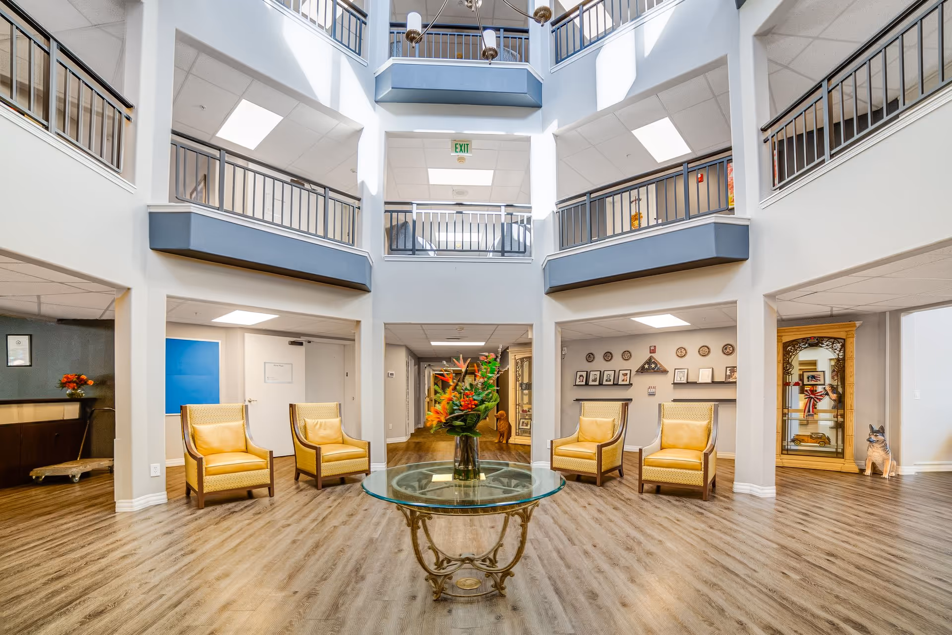 Spacious and well-lit interior common area with high ceilings and a second-floor balcony overlooking the space. The room features four yellow armchairs arranged around a round glass table with a floral centerpiece. The floor is wood-style laminate, and the walls are painted white. There are framed pictures and decorative items on shelves and a cabinet visible in the background.