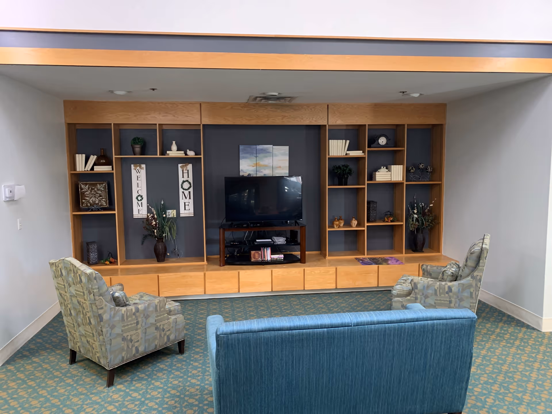 A cozy living room area with a blue couch and two patterned armchairs facing a wooden entertainment center with shelves. The shelves hold decorative items, books, plants, and a flat-screen TV. The wall behind the entertainment center is painted dark gray, and there are two vertical signs that say 'WELCOME' and 'HOME'. The floor is carpeted with a patterned design.