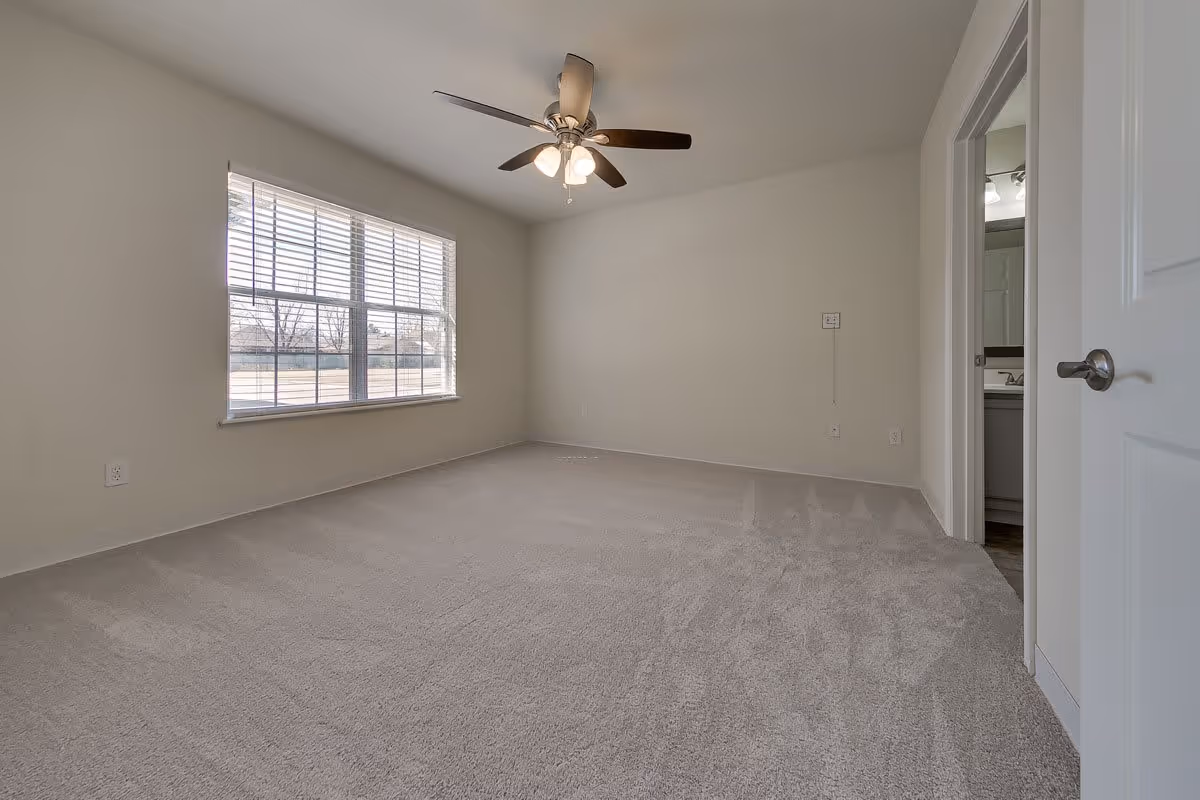 Empty bedroom with beige carpet, a large window with blinds letting in natural light, a ceiling fan with lights, and a partially open door revealing a bathroom sink and mirror.