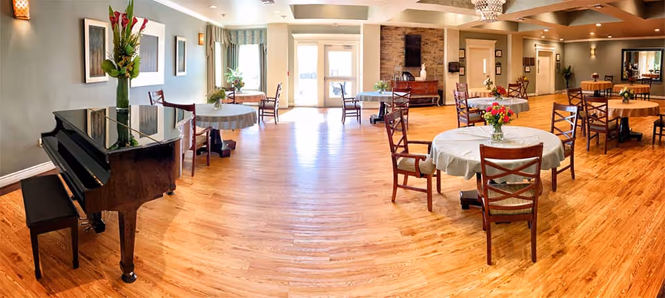 A spacious, well-lit dining area with round tables covered in white tablecloths, each adorned with a small vase of flowers. Wooden chairs surround the tables. A black grand piano with a floral arrangement on top is positioned on the left side of the room. The room has wooden flooring, soft wall lighting, and large windows with curtains allowing natural light to enter.
