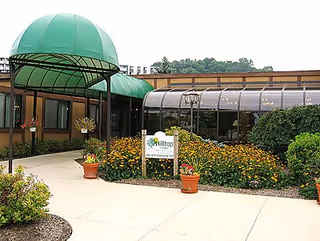 Entrance to Hilltop Village facility with a green canopy over the walkway, surrounded by flower beds and shrubs. A sign near the entrance reads 'Hilltop Village Reception Center.'