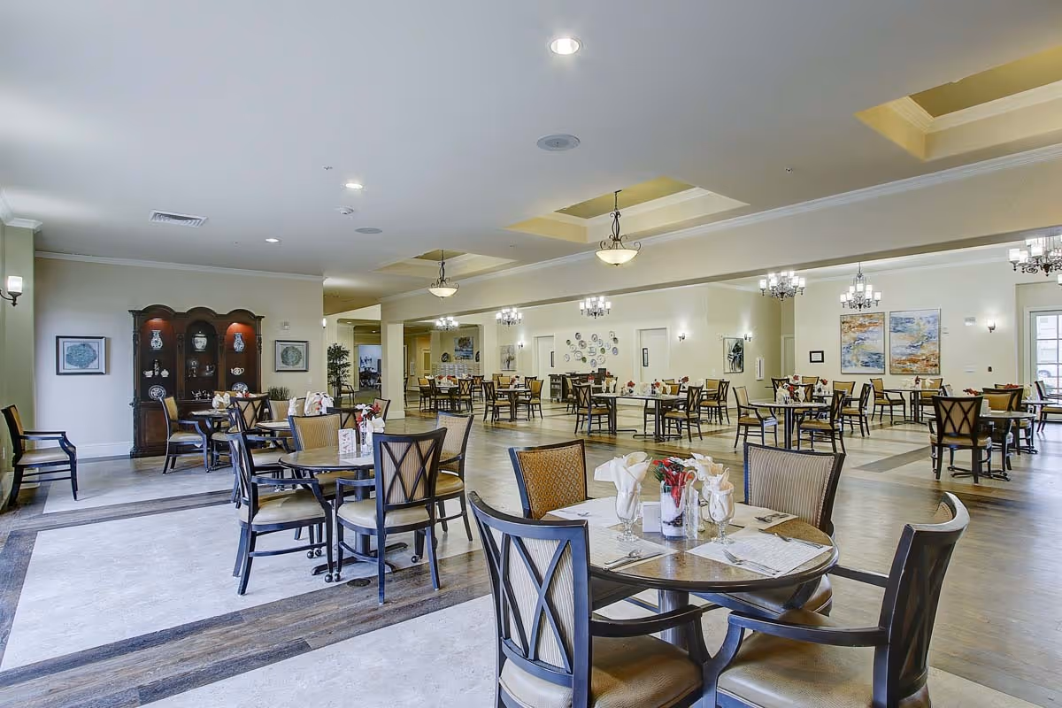 A spacious and well-lit dining room in a senior living facility with multiple round tables and chairs arranged neatly. Each table is set with napkins, glasses, and small floral centerpieces. The room features elegant chandeliers, wall sconces, framed artwork, and a wooden cabinet displaying decorative items.