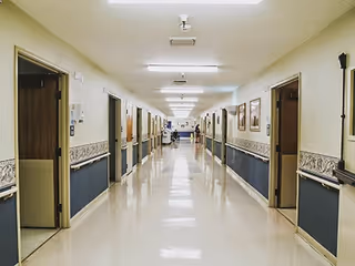 A long, clean hallway in a nursing and rehabilitation center with multiple open doors on both sides, beige walls with a blue lower section and decorative border, and fluorescent ceiling lights.