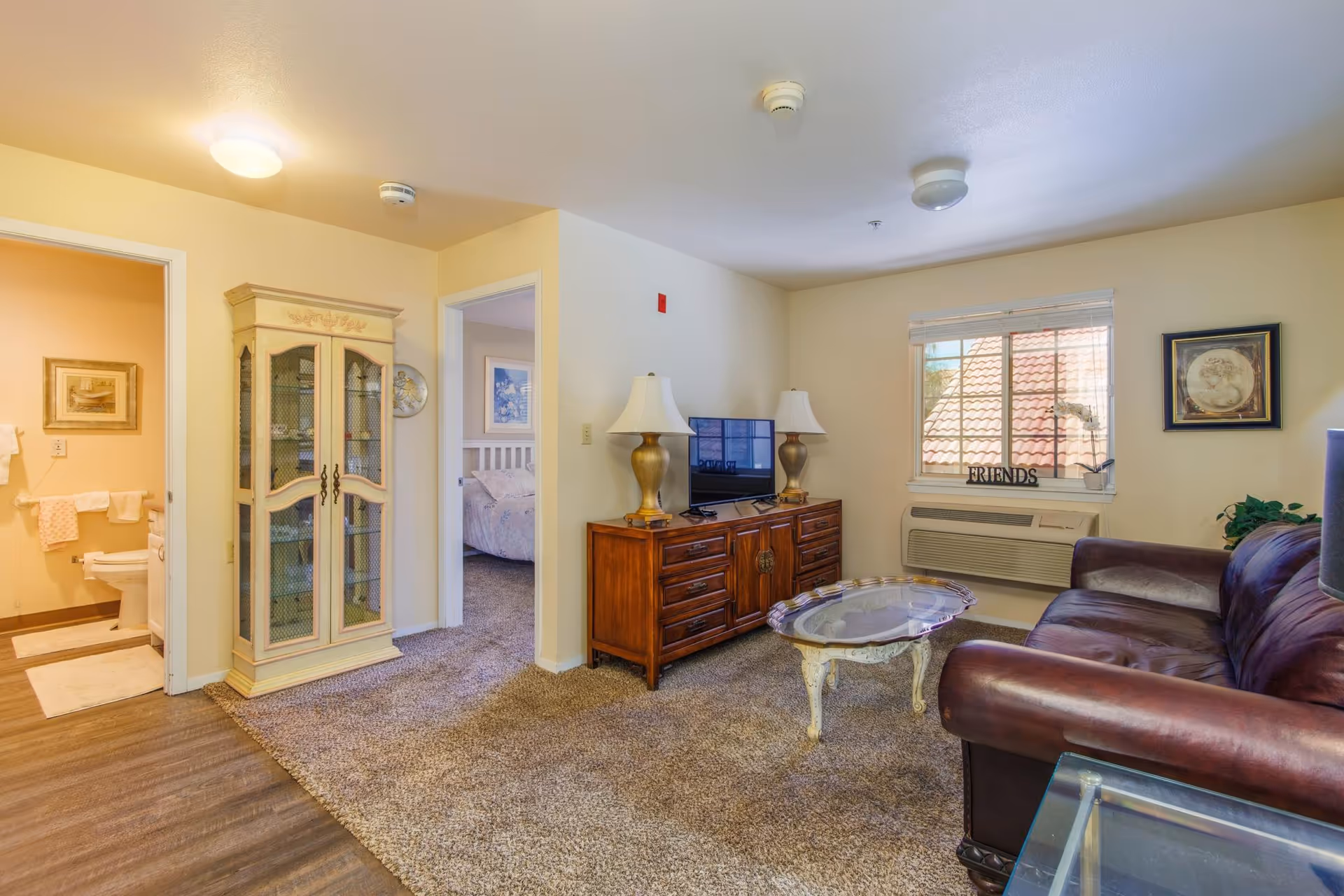 Cozy living room in an assisted living unit with a leather sofa, glass-top coffee table, TV on a wooden cabinet, and doorways to a bedroom and bathroom.