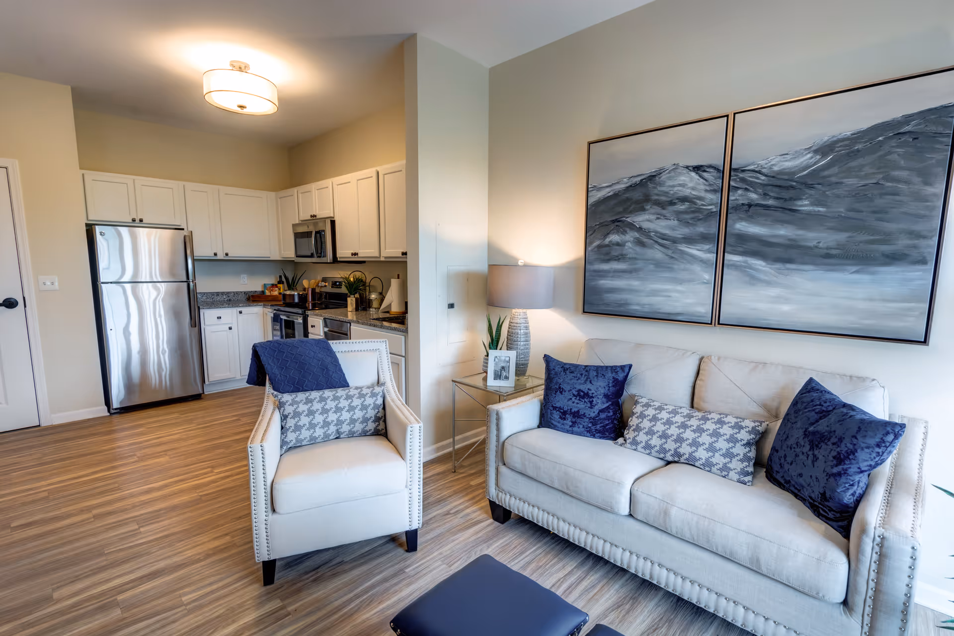 A modern senior living apartment interior featuring a small kitchen with white cabinets, stainless steel refrigerator, microwave, and stove. Adjacent to the kitchen is a cozy living area with a beige sofa adorned with blue and patterned pillows, a matching armchair with a patterned pillow and blue throw, a side table with a lamp and framed photo, and two large abstract paintings on the wall.