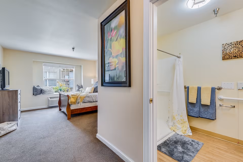 View of a senior living facility room showing a bedroom with a wooden bed, a dresser with a TV, a window with cushions on the sill, and a bathroom with a shower, towels hanging on the wall, and a bath mat on the floor.