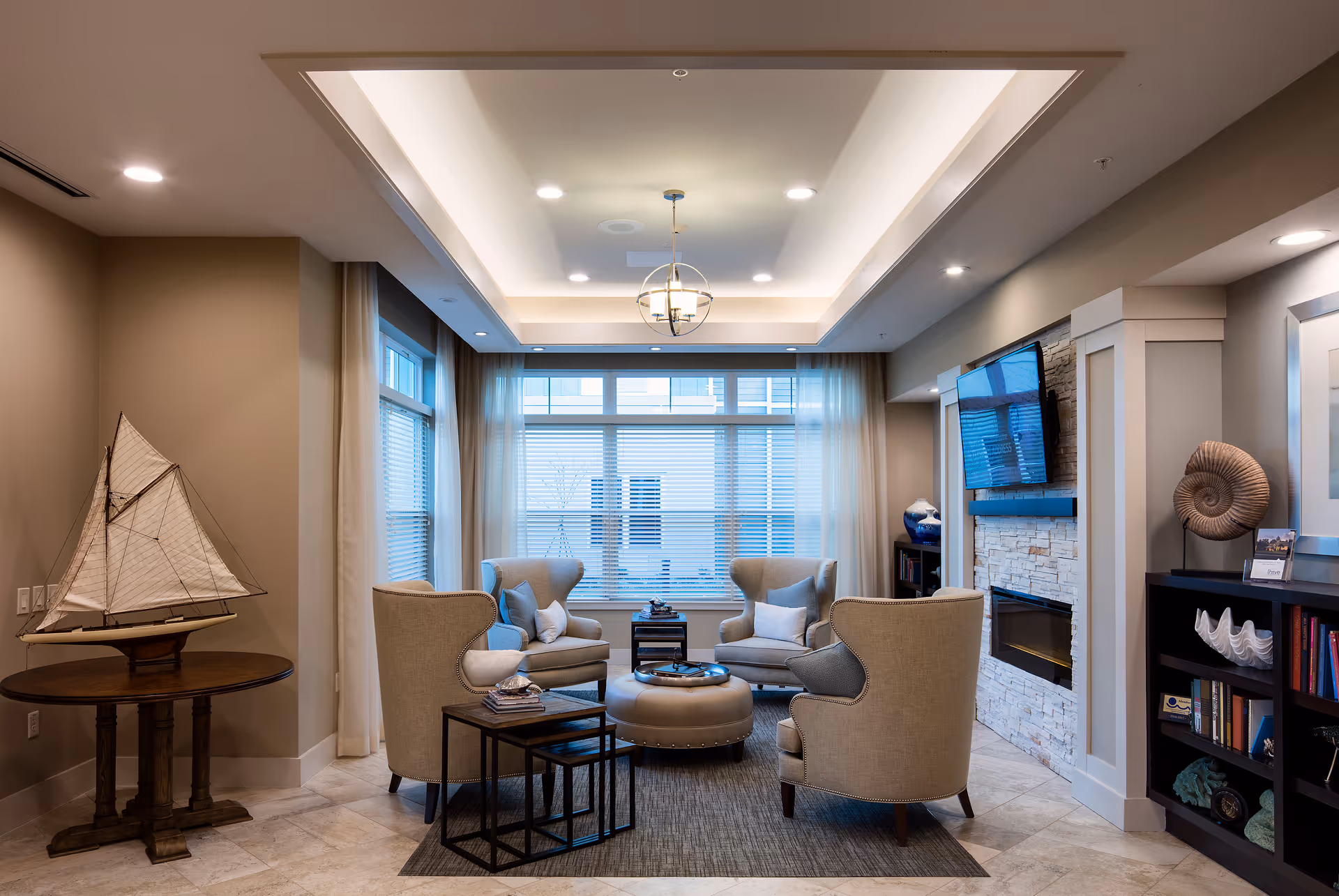A cozy living room area with four beige armchairs arranged around a round ottoman on a rug. There is a small side table with nested tables next to one chair. A large window with blinds and sheer curtains lets in natural light. A flat-screen TV is mounted above a modern electric fireplace with a stone surround. Decorative items include a model sailboat on a round wooden table, a large shell sculpture on a bookshelf, and various books and decor pieces on shelves.