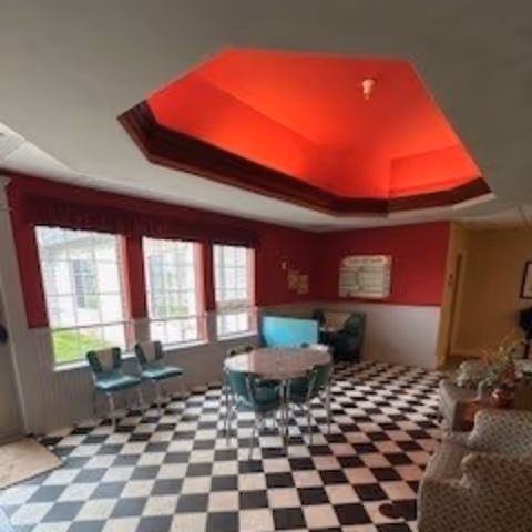 Interior view of a common area with black and white checkered flooring, a round table with green chairs, a red ceiling with recessed lighting, large windows with red valances, and additional seating including armchairs and a small couch.