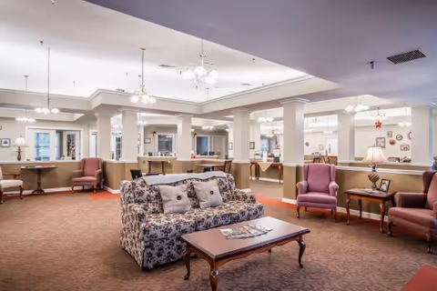 A spacious senior living facility common area with floral patterned sofa, several armchairs, wooden coffee tables, and soft lighting from ceiling fixtures. The room has beige walls with white trim and carpeted floors, creating a warm and inviting atmosphere.
