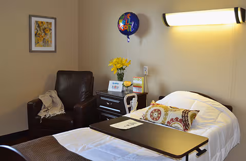 A nursing facility room with a hospital bed covered in white linens and decorative pillows, a dark brown leather armchair with a beige throw blanket, a bedside table with a vase of yellow flowers, a 'Get Well Soon' balloon, and a framed picture on the wall.