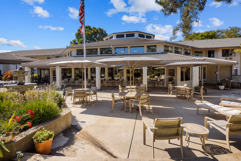 Outdoor patio area at Provincial Chico Senior Living with several tables and chairs under large umbrellas, surrounded by greenery and a water fountain, with the senior living building in the background under a blue sky with some clouds.