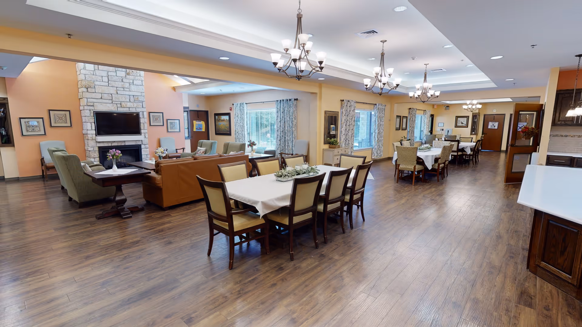 Spacious senior living common area with multiple dining tables covered with white tablecloths and surrounded by chairs. The room features wooden flooring, several chandeliers, large windows with curtains, and a seating area with armchairs and a sofa near a stone fireplace with a mounted TV above it.
