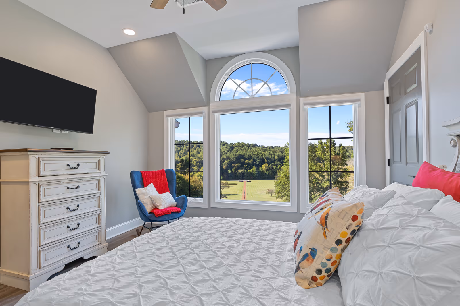 A bright bedroom with a large bed covered in white bedding and colorful pillows. There is a blue armchair with a red throw blanket and white pillows near three large windows, including an arched window, showing a scenic view of green trees and a clear blue sky. A white dresser with a flat-screen TV mounted above it is on the left side of the room.