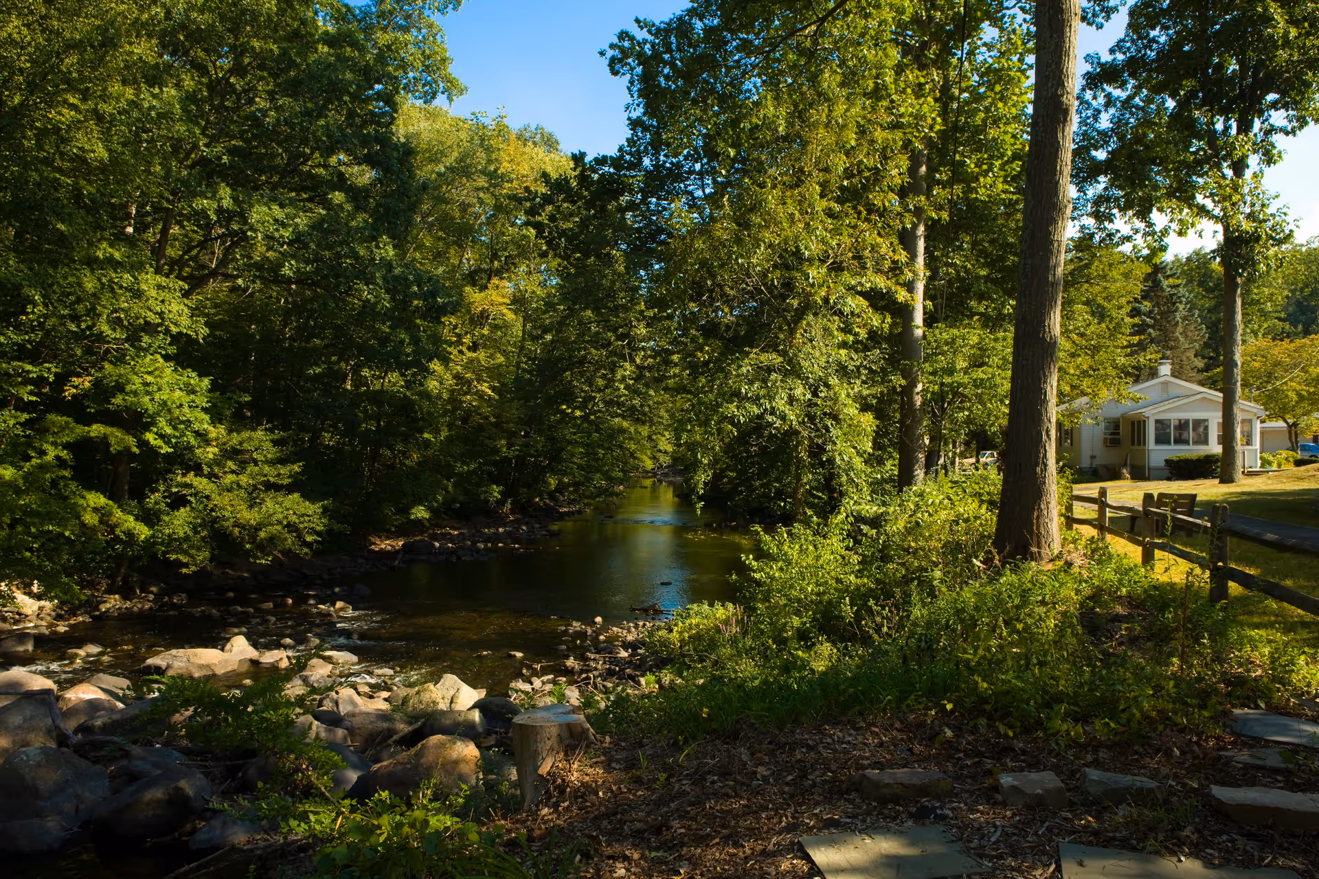 A peaceful outdoor scene featuring a gently flowing river surrounded by lush green trees and rocks, with a small white building and a wooden fence visible on the right side under a clear blue sky.