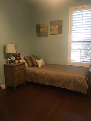Small single bedroom with a bed against a mint-green wall, a nightstand with a lamp and teddy bear, and a window with white shutters.