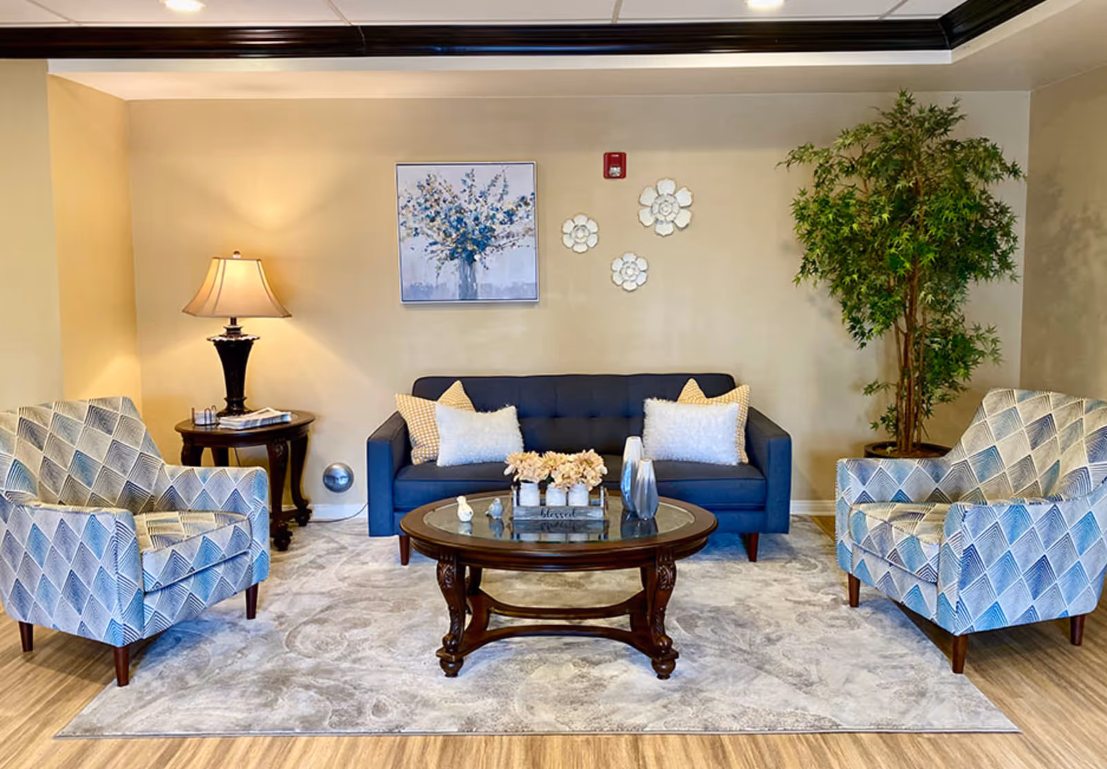 A cozy living room with a blue sofa adorned with white and yellow pillows, two patterned armchairs, a round wooden coffee table with decorative items, a side table with a lamp, a large potted plant, and wall decorations including a floral painting and three white flower-shaped ornaments.
