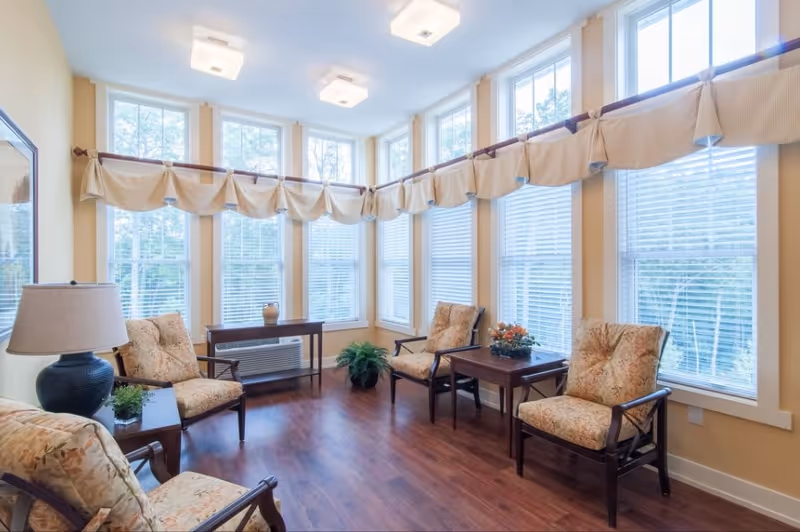 A bright and cozy sitting area with multiple large windows letting in natural light. The room features four cushioned armchairs with floral upholstery arranged around two wooden side tables. There is a table lamp on one side and decorative plants on the tables and floor. The walls are painted a soft yellow, and the floor is a polished wood finish.