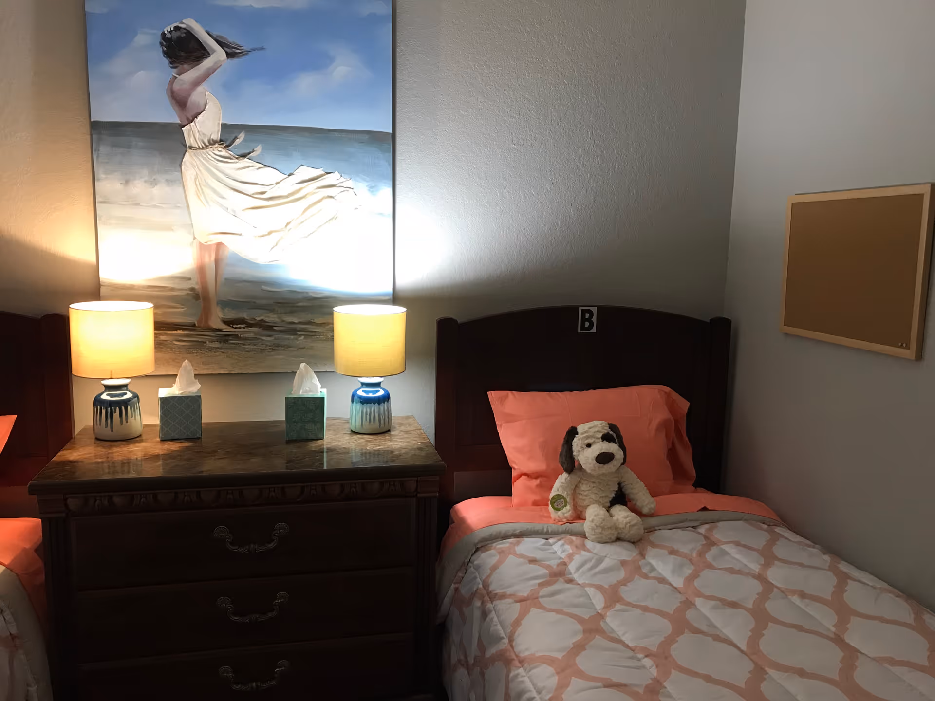 A cozy bedroom with a single bed labeled with the letter B. The bed has coral-colored sheets and a patterned comforter, with a stuffed dog toy sitting on the pillow. Next to the bed is a wooden nightstand with two lit lamps and two tissue boxes. Above the nightstand hangs a painting of a woman in a white dress standing on a beach with the ocean and sky in the background. There is also a corkboard on the adjacent wall.