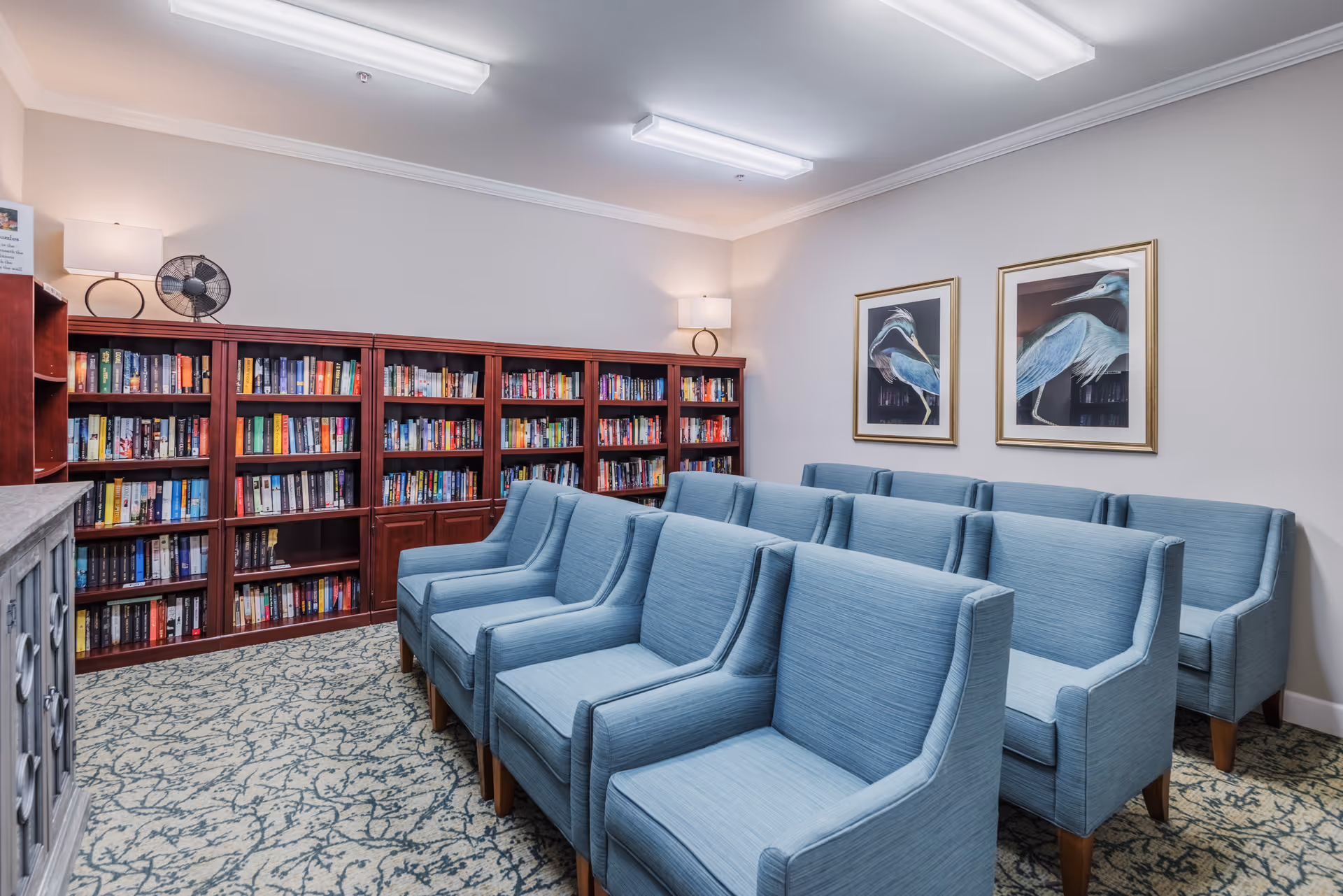 A small library/reading room with rows of blue upholstered chairs, wooden bookshelves filled with books, and framed bird prints on the wall.