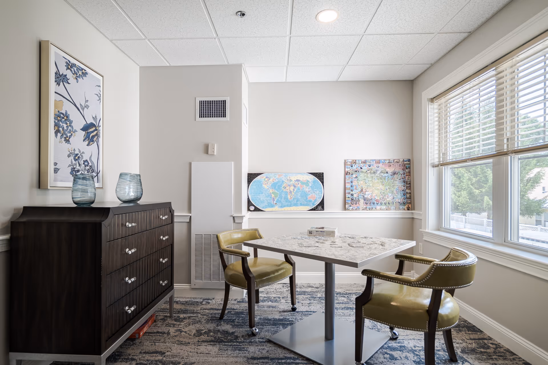 A small, well-lit room with a square table and two green upholstered chairs on wheels. There is a dark wooden dresser with two blue glass vases on top and a framed floral artwork hanging above it. On the far wall, there are two maps displayed on a ledge. A large window with blinds allows natural light to enter the room.